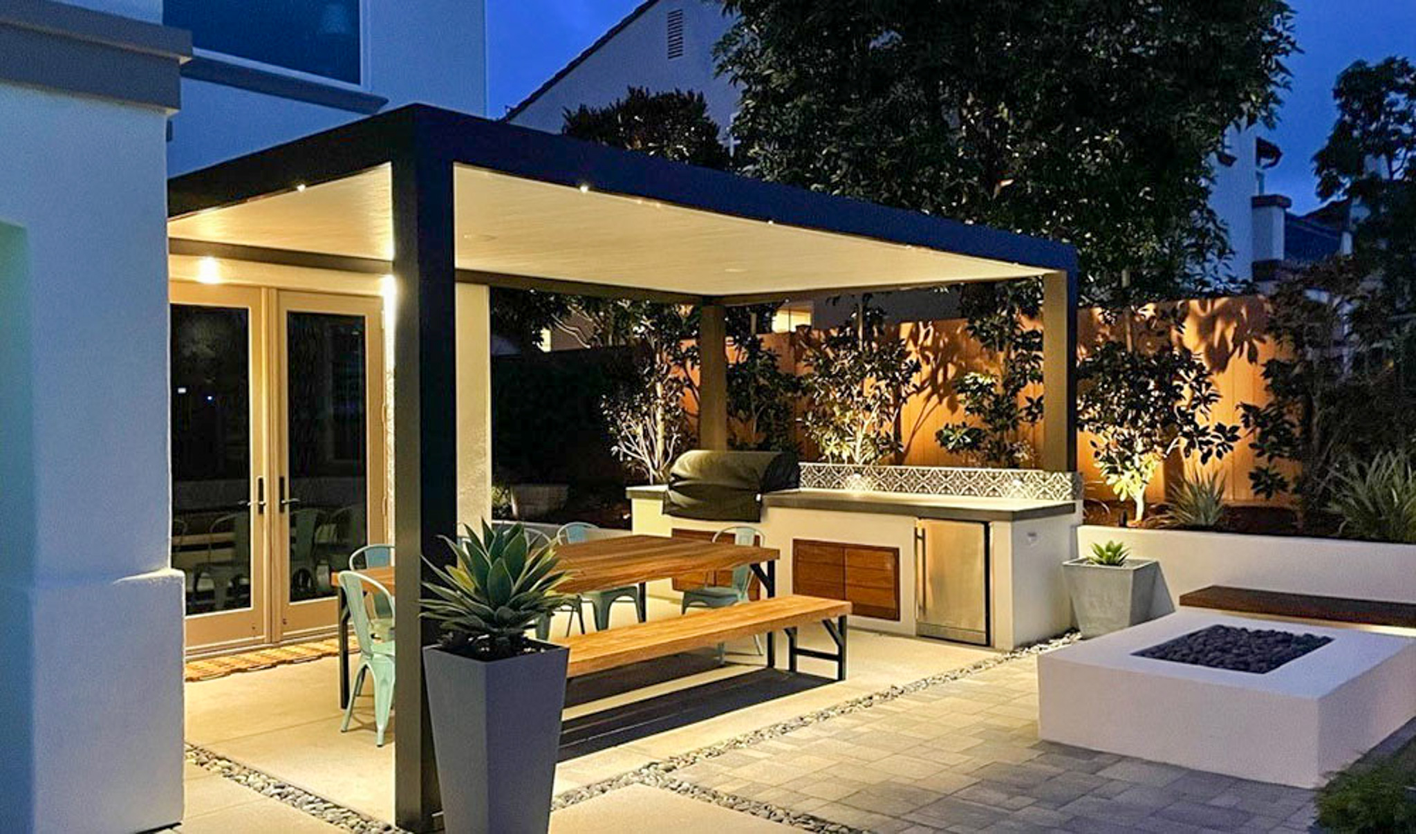 Contemporary style solid patio cover made of aluminum over outdoor sitting area with table and barbecue, at night with lights on in outdoor space