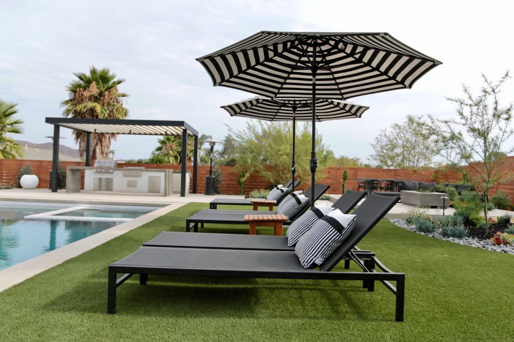View of a backyard in San Diego California with black and white furniture on turf next to a pool and outdoor kitchen covered by a contemporary solid freestanding patio cover