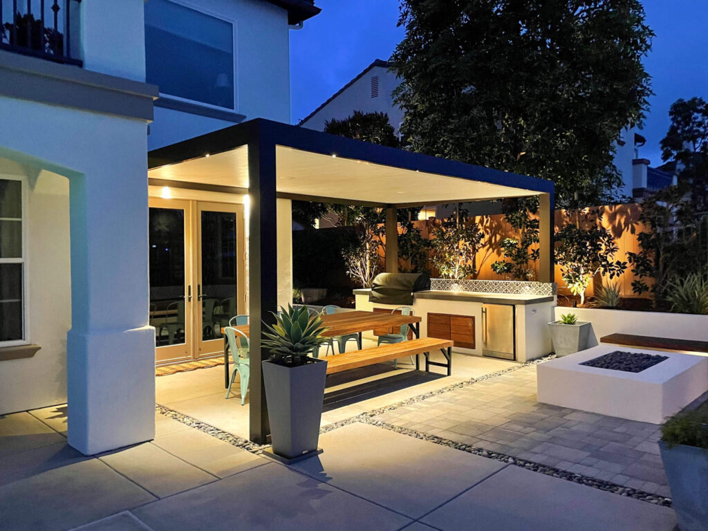Contemporary style solid patio cover made of aluminum over outdoor sitting area with table and barbecue, at night with lights on in outdoor space