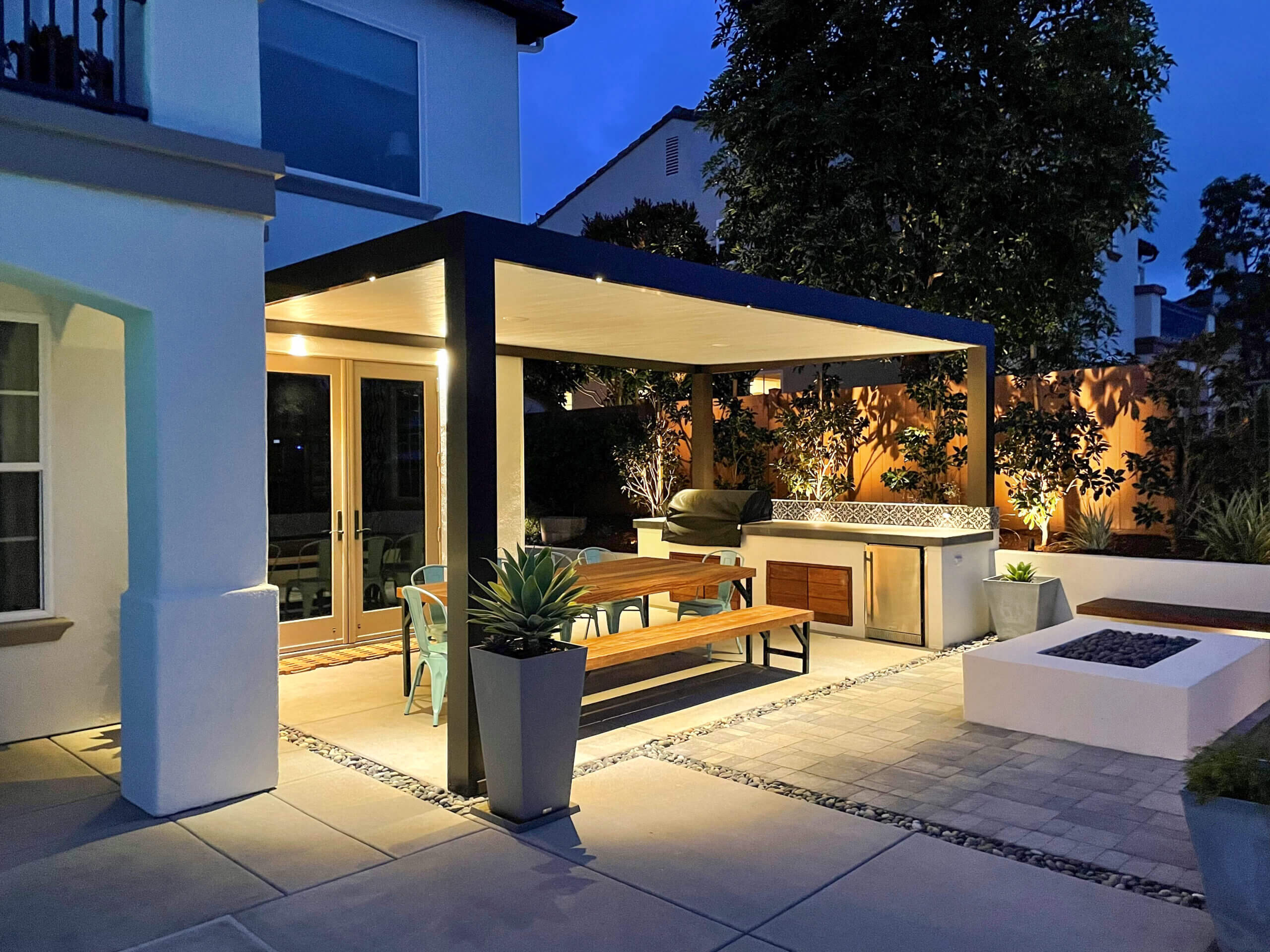 Contemporary style solid patio cover made of aluminum over outdoor sitting area with table and barbecue, at night with lights on in outdoor space
